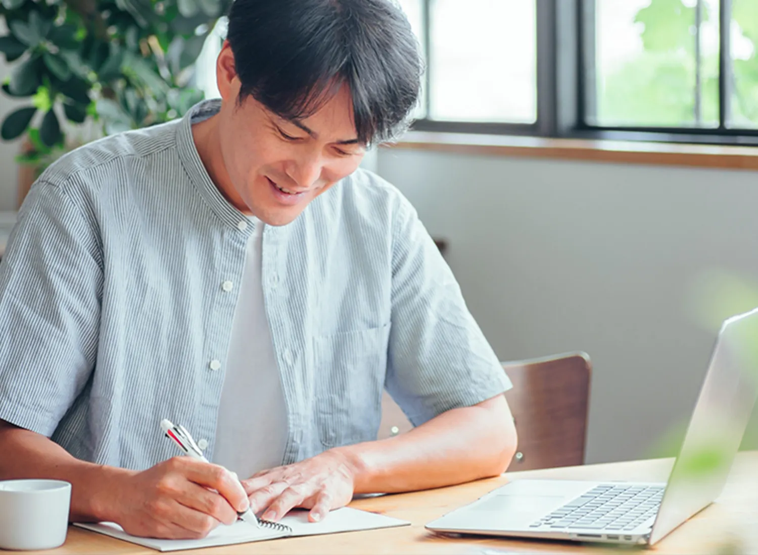 ノートパソコンを前に置いて、笑顔でノートにボールペンで書き込んでいる男性の様子